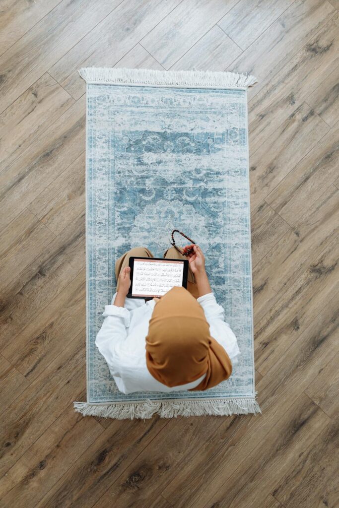 Overhead view of a Muslim woman praying with a tablet displaying scripture and holding prayer beads.