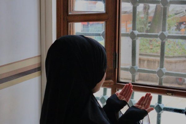A serene image of a woman in a headscarf praying by a window, symbolizing devotion and spirituality.